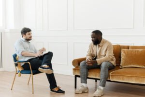 A professional Black man sitting on a sofa talking to a men's PTSD therapist. PTSD counseling in Portland, OR can help you heal from traumatic memories. Get the personalized support you deserve today. 