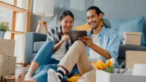 A happy couple sitting next to moving boxes while looking at a tablet. An anxiety therapists in Portland, OR can help you work through relationship anxiety. If you want to get started, call us today!