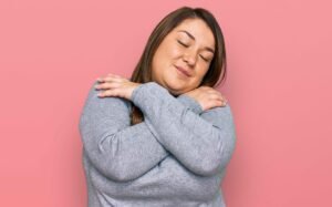 A woman hugging herself, showing self-love in front of a pink background. This represents how our relationship anxiety therapy in Portland, OR can help you with insecurity & jealousy. Our anxiety therapists are here to help!