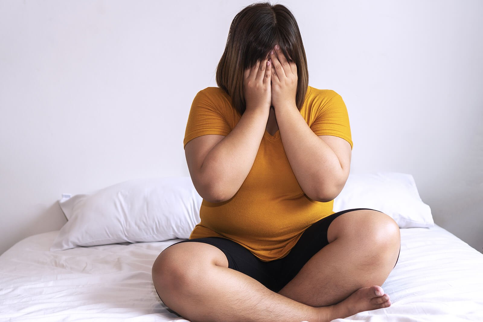 A woman sitting cross-legged on a white bed while covering her eyes with her hands. Representing how sleep can affect your anxiety & every day life. Anxiety therapy in Portland, OR can help you develop a sleep routine that works!