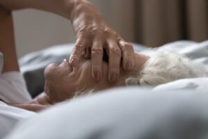 An elderly woman covering her eyes with her hands while laying in bed. 