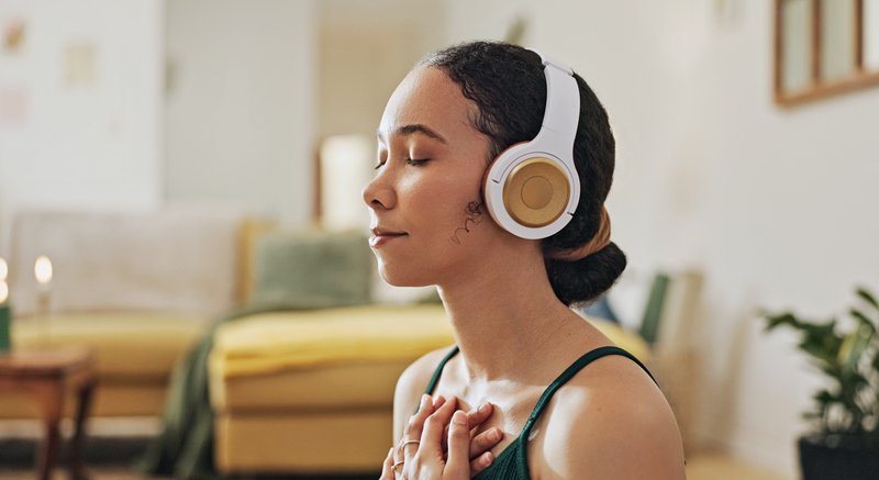 A woman with eyes closed and hands on her chest, wearing headphones in a calm home setting—practicing mindfulness and emotional awareness.