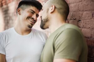 A biracial LGBTQ+ couple smiling at each other in front of a brick wall. Trauma counseling in Portland, OR can help you reclaim your life. Start healing today!